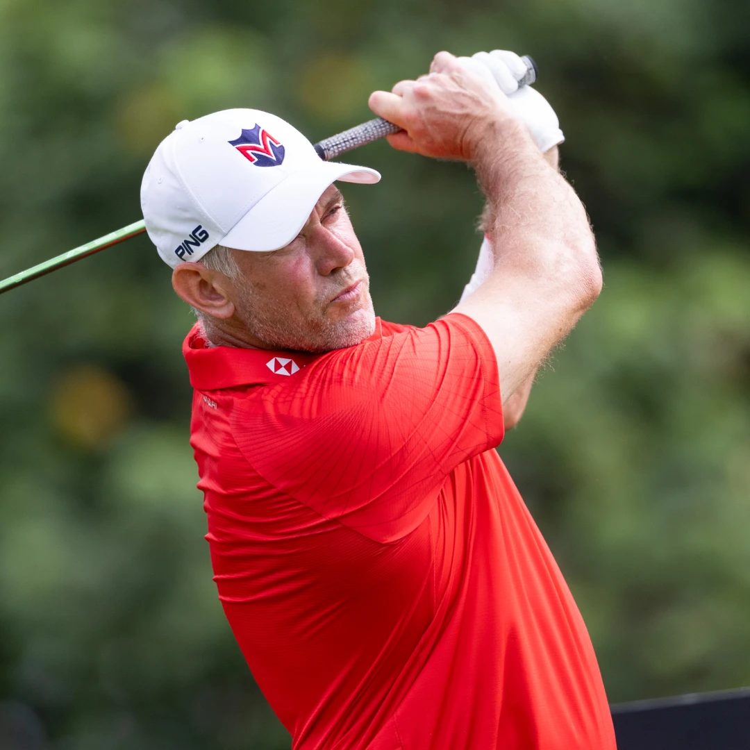 Man in a red shirt and white cap swings a golf club, focused gaze, with greenery in the background.