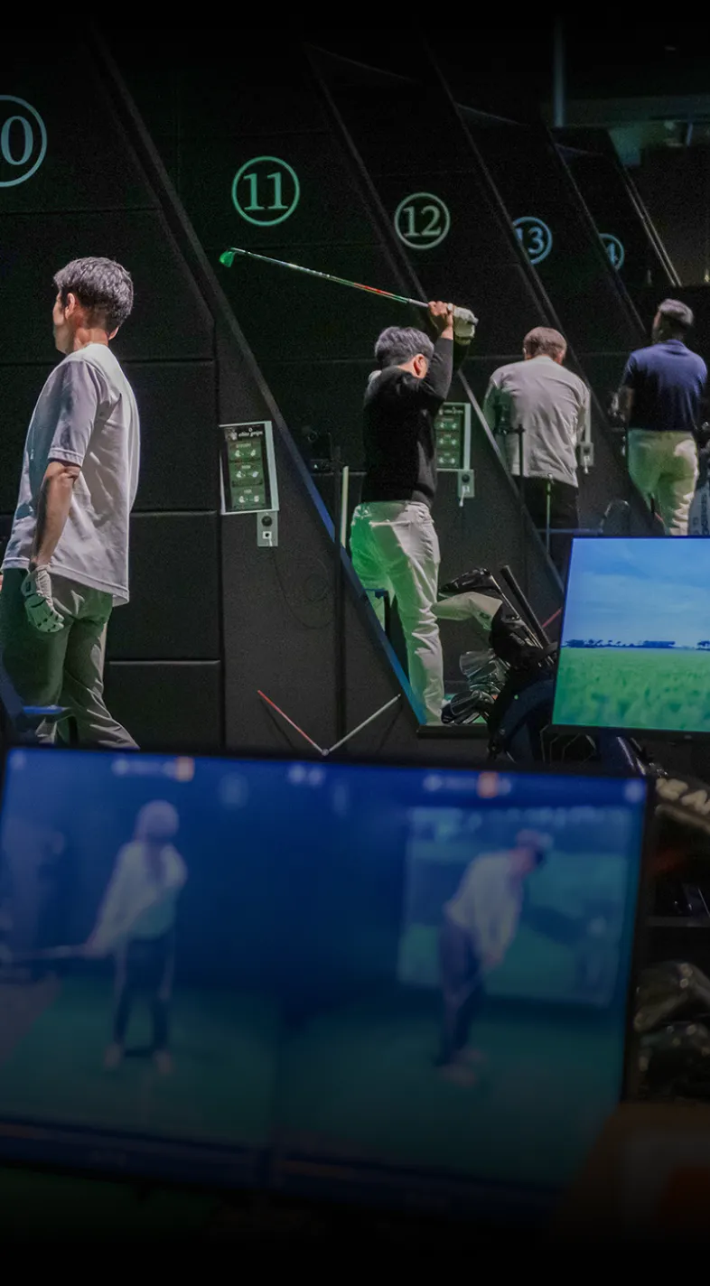 People practicing golf swings in an indoor facility with numbered stalls and screens displaying their movements.