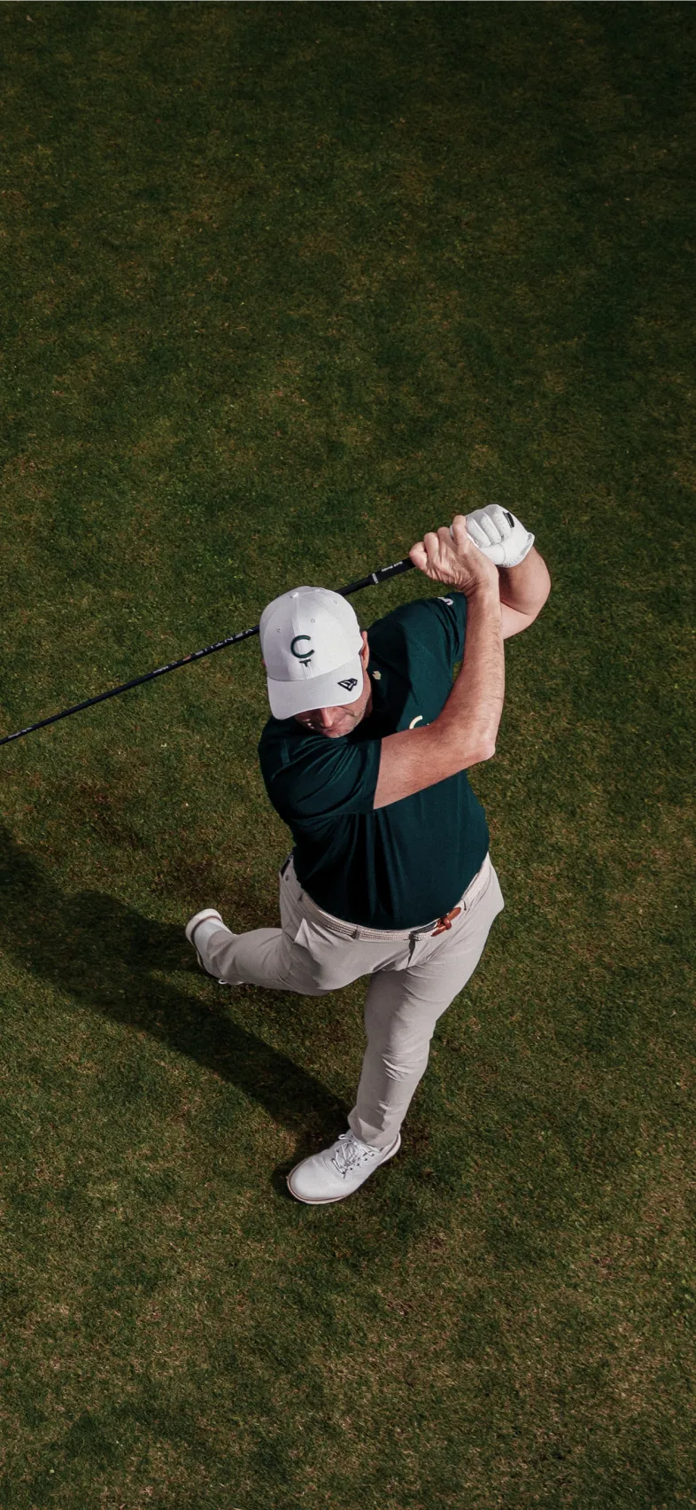Golfer in white cap and gloves swinging a club on the green, captured from above.