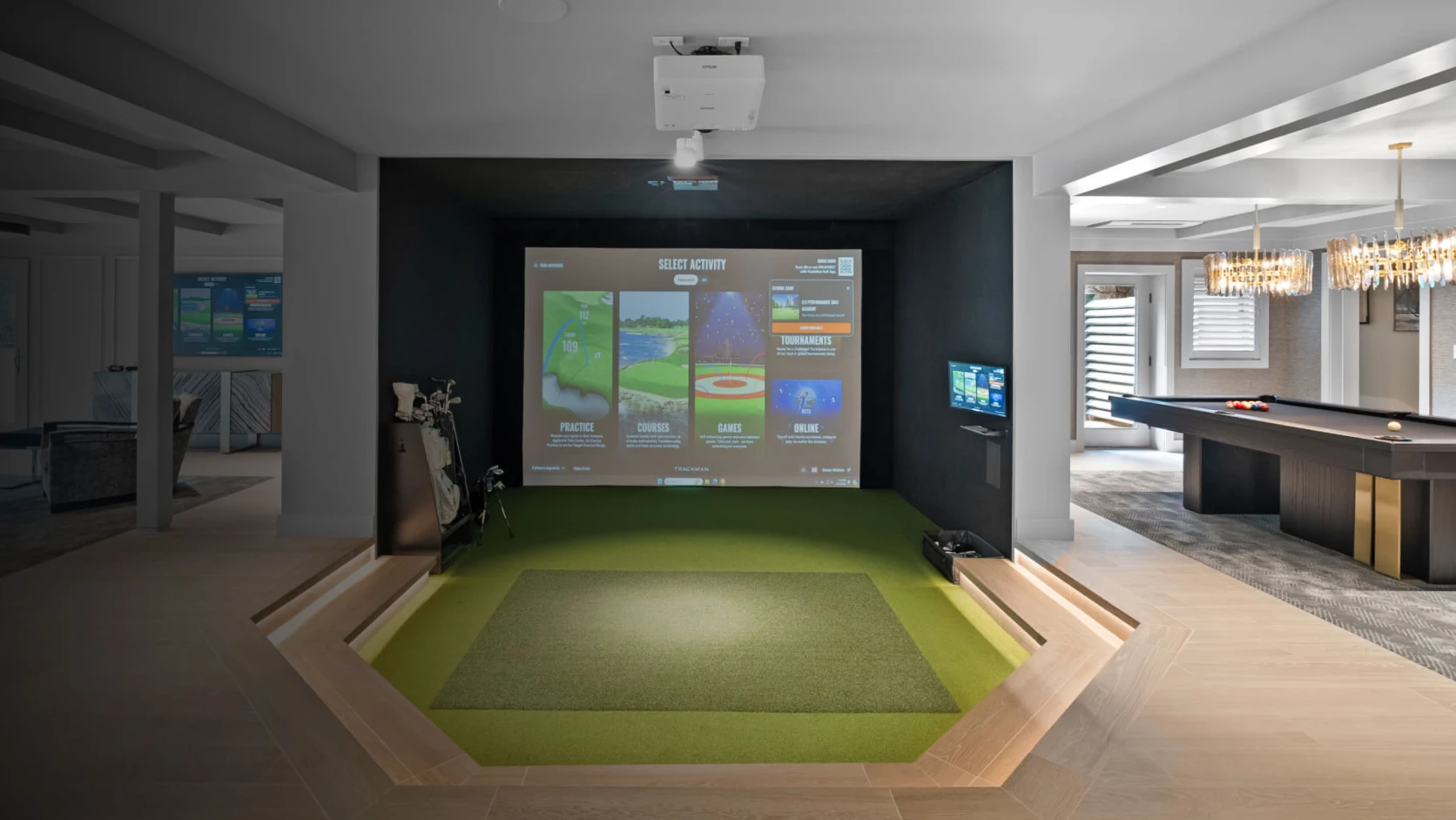 Sunken indoor golf simulator with projector screen, turf hitting mat and clubs in a modern basement game room with a pool table.