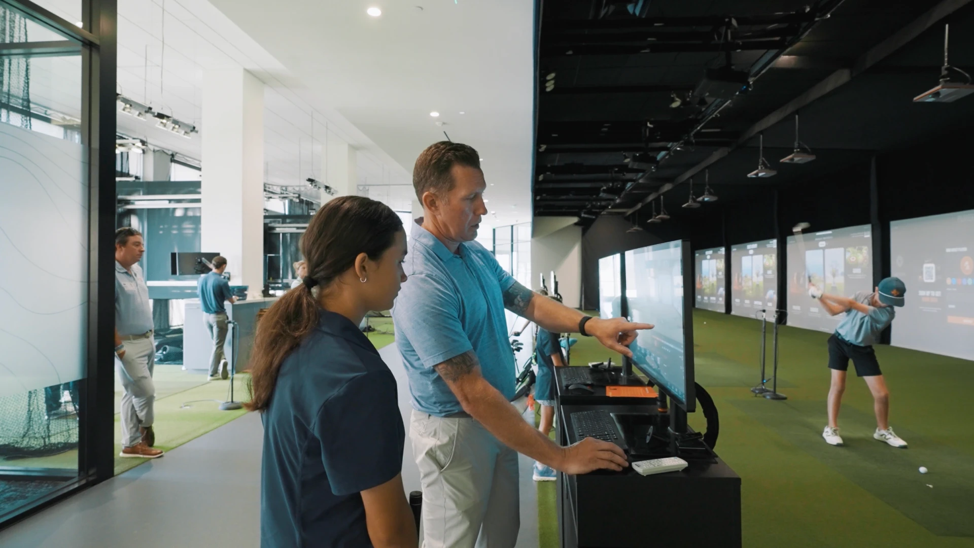 A man and a woman are looking at a monitor in a golf simulation facility, while a person swings a golf club in the background.