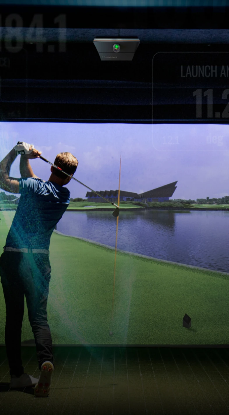 Person swinging a golf club indoors, facing a large screen displaying a golf course with water and modern architecture.